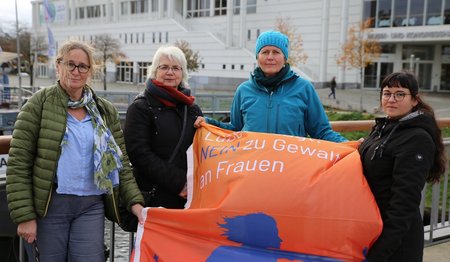 Vier Frauen stehen mit einer orangefarbenen Fahne vor der Lübecker Musik- und Kongresshalle.