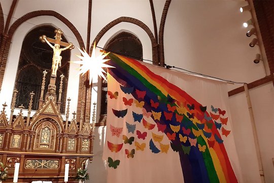 Ein Altar mit Jesus am Kreuz im Hintergrund, einem hell strahlenden gelben Stern und im Vordergrund eine Leinwand mit einem bunten Regenbogen und vielen bunten kleinen Schmetterlingen, welche gen Himmel, nach oben fliegen