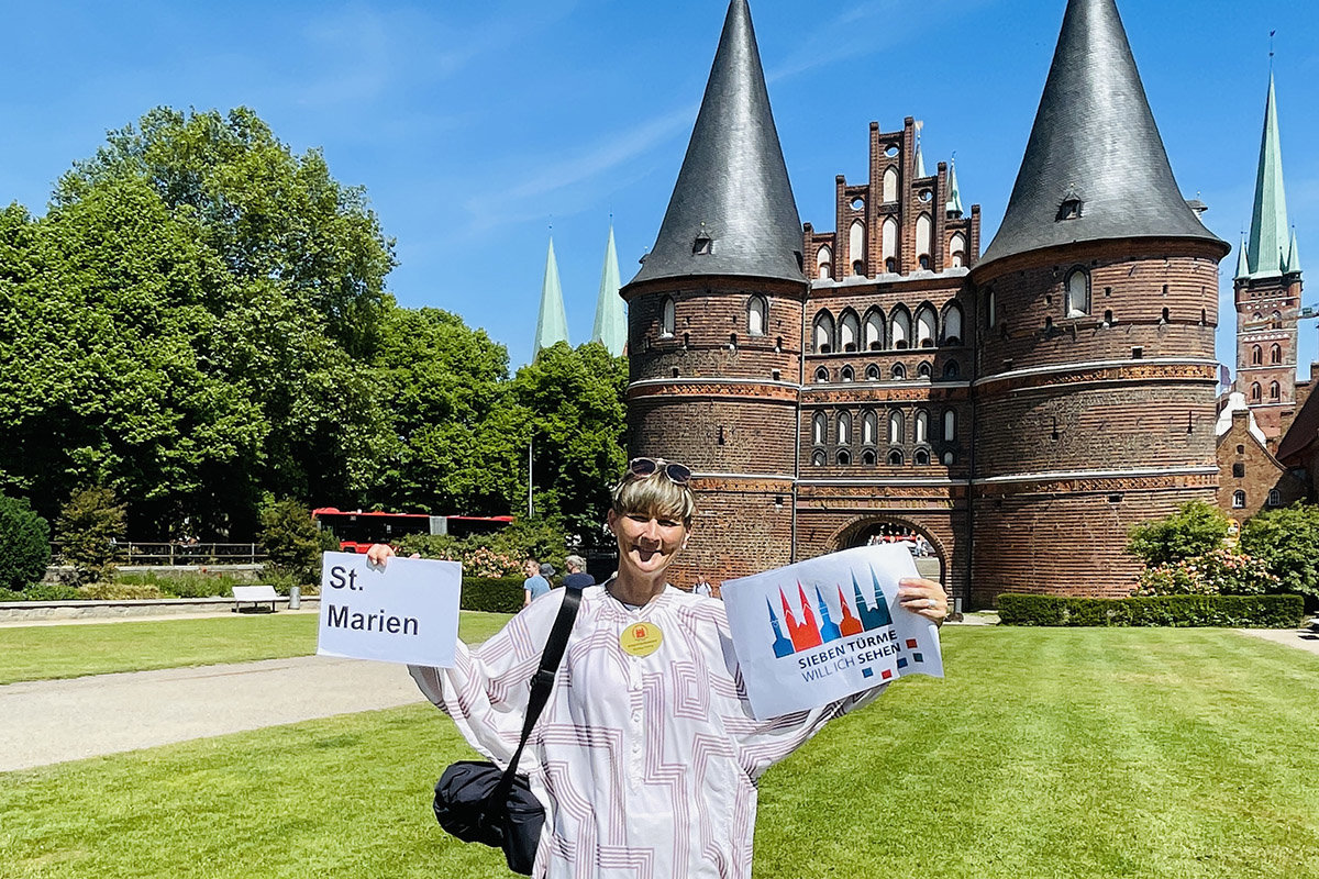 Frau Kramp hält in der linken Hand ein Schild auf welchem St. Marien steht, in der rechten das Logo vom Sieben-Türme-Projekt. Im Hintergrund ist das Holstentor und rechts daneben St. Petri zu sehen. Ein klarer blauer Himmel und Sonnenschein
