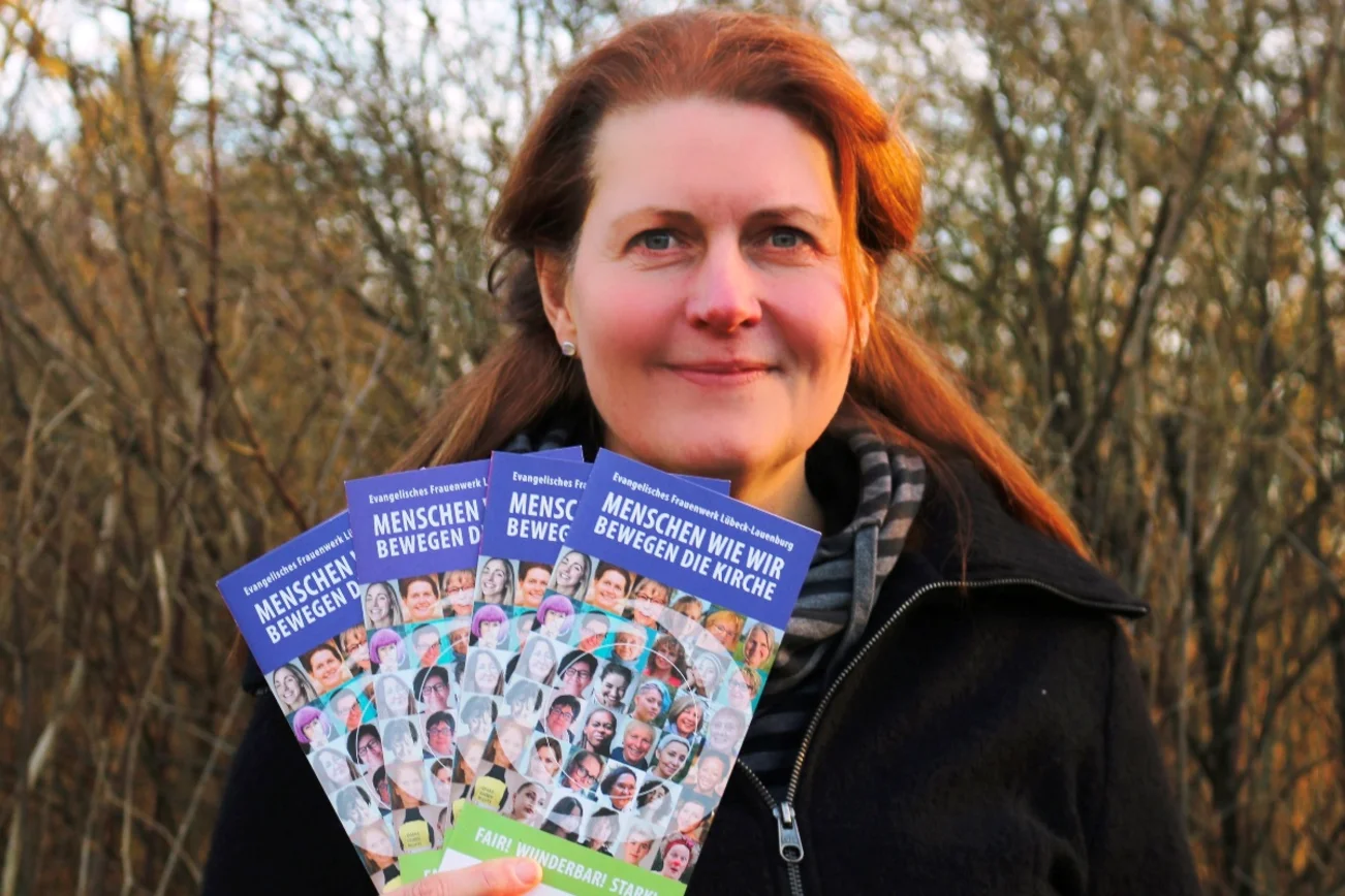 Silke Meyer blickt in die Kamera. Sie hält Flyer des Frauenwerks aufgefächert in der Hand. Im Hintergrund ist eine Hecke.