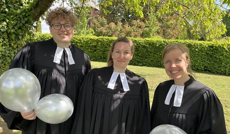Ein Mann und zwei Frauen stehen im Talar in einem Garten und halten Luftballons.