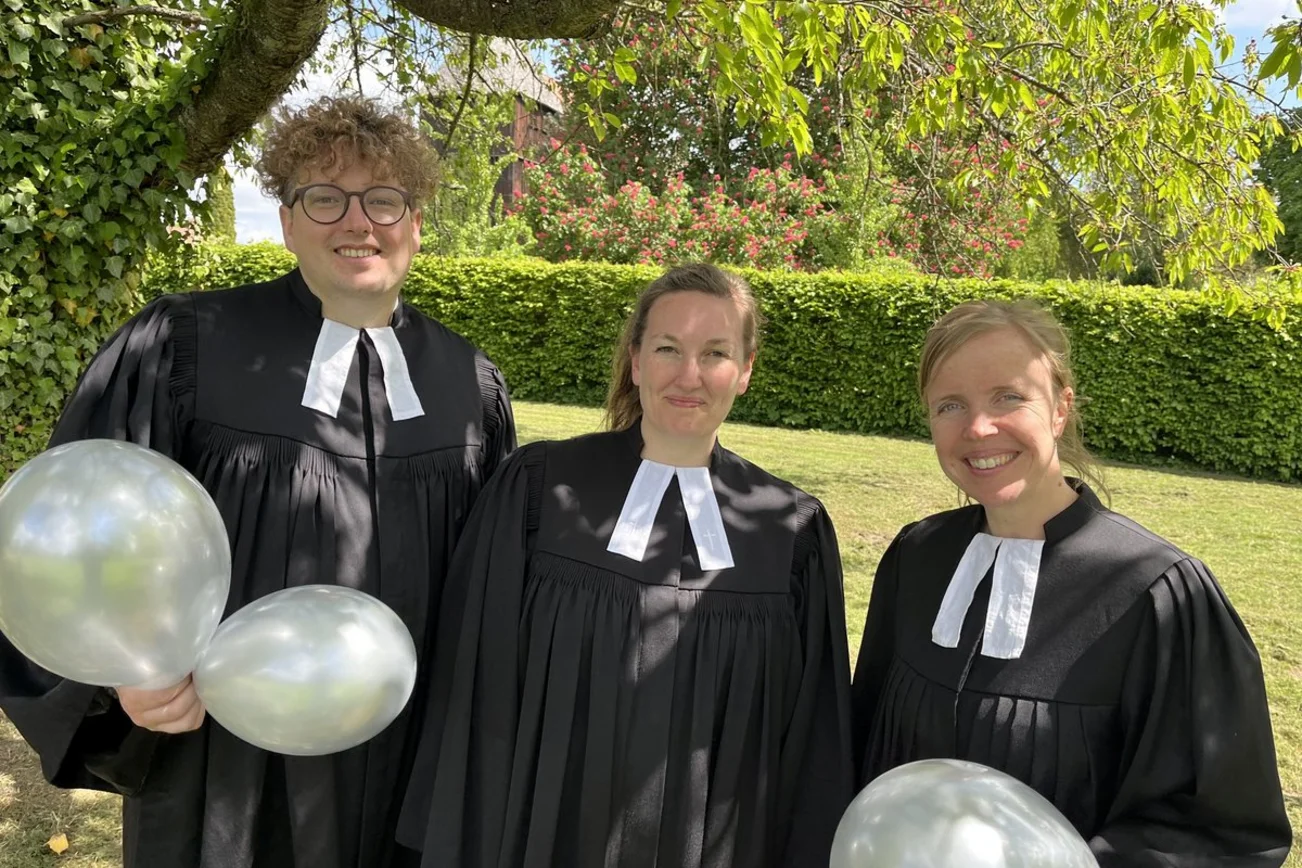 Ein Mann und zwei Frauen stehen im Talar in einem Garten und halten Luftballons.