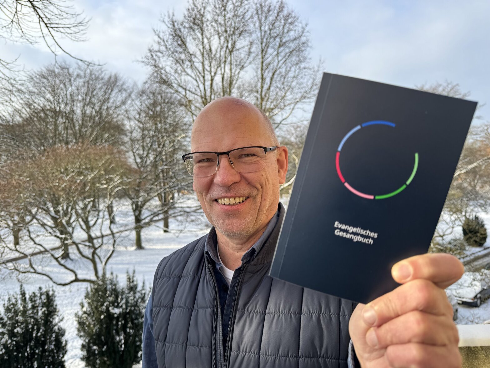Ein Mann steht in einem verschneiten Park und hält lächelnd ein Buch in die Kamera.
