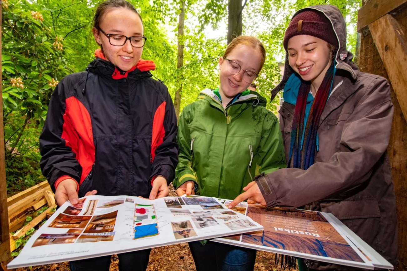Drei Mädchen stehen im Wald und schauen auf Baupläne. 