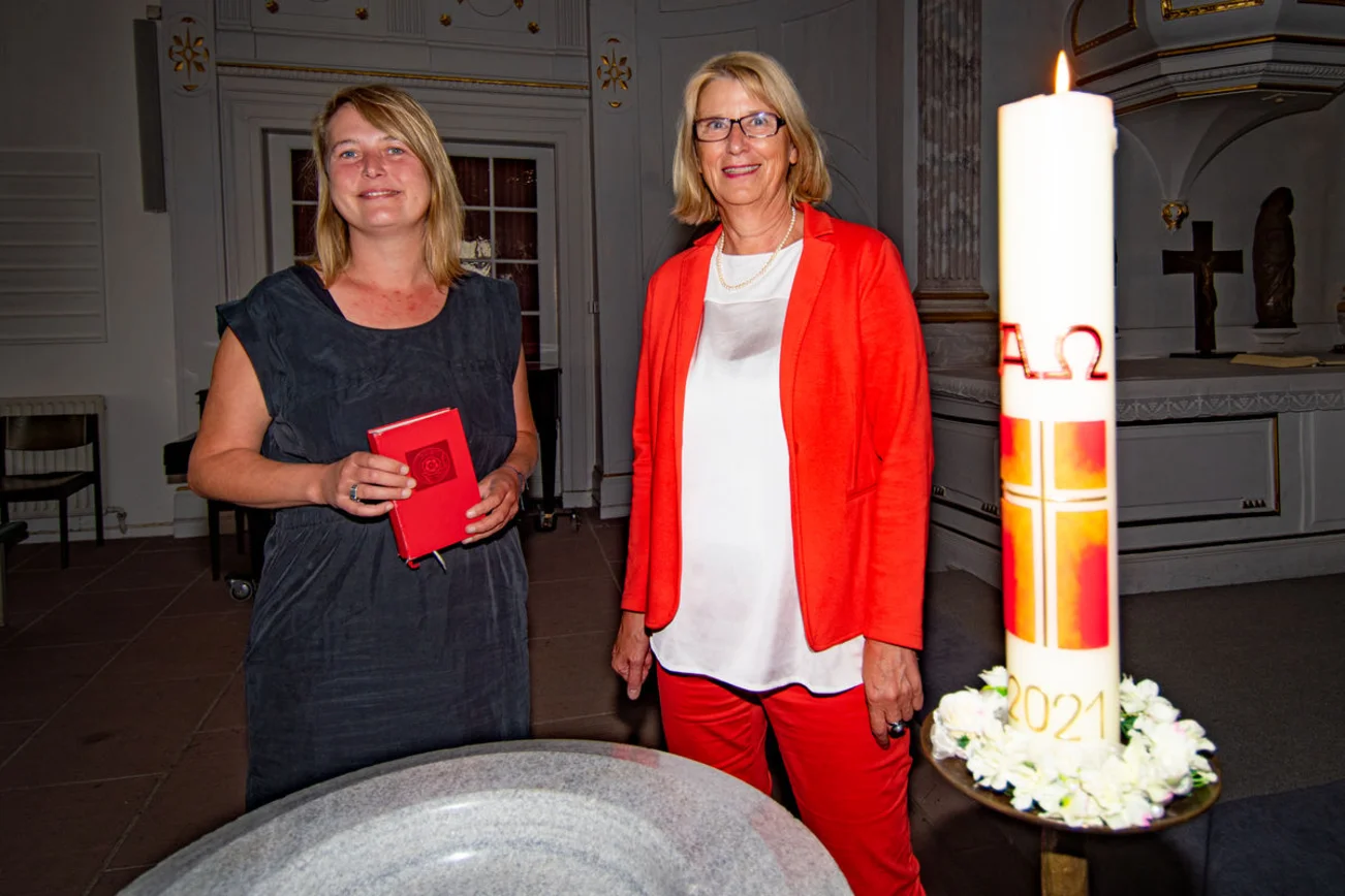 Zwei Frauen stehen an einem Taufbecken neben einer Kerze in einer Kirche.
