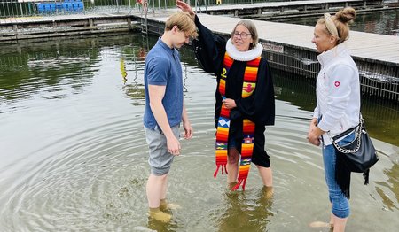 Pastorin Nicola Nehmzow tauft Ben Joel Hinz. Mit dabei: Mutter Kati Hinz. Sie stehen alle im Wasser, die Pastorin hält eine Hand über den Kopf des jungen Mannes.