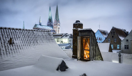 Abenddämmerung mit schneebedeckten Dächern von Lübeck, in der Mitte eine offene Dachluke aus der Licht scheint, im Hintergrund St. Marien