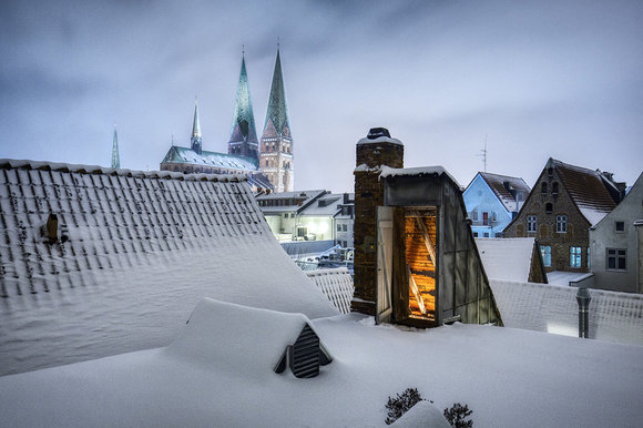 Abenddämmerung mit schneebedeckten Dächern von Lübeck, in der Mitte eine offene Dachluke aus der Licht scheint, im Hintergrund St. Marien