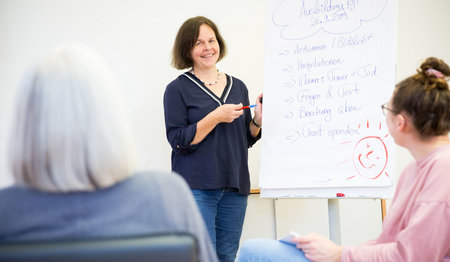 Frau steht neben einem Flipchart und erklärt, zwei Personen sind von hinten zu sehen.