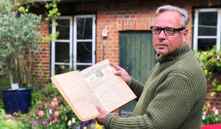 Ein Mann hält ein altes Buch aufgeschlagen in der Hand. Er zeigt die alten Seiten in Richtung Kamera. Im Hintergrund Teile eines Hauses aus Backstein mit grüner Holztür und weißen Sprossenfenstern. Frühlingsblumen blühen davor.