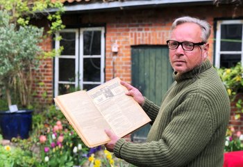 Ein Mann hält ein altes Buch aufgeschlagen in der Hand. Er zeigt die alten Seiten in Richtung Kamera. Im Hintergrund Teile eines Hauses aus Backstein mit grüner Holztür und weißen Sprossenfenstern. Frühlingsblumen blühen davor. - Copyright: Ines Langhorst