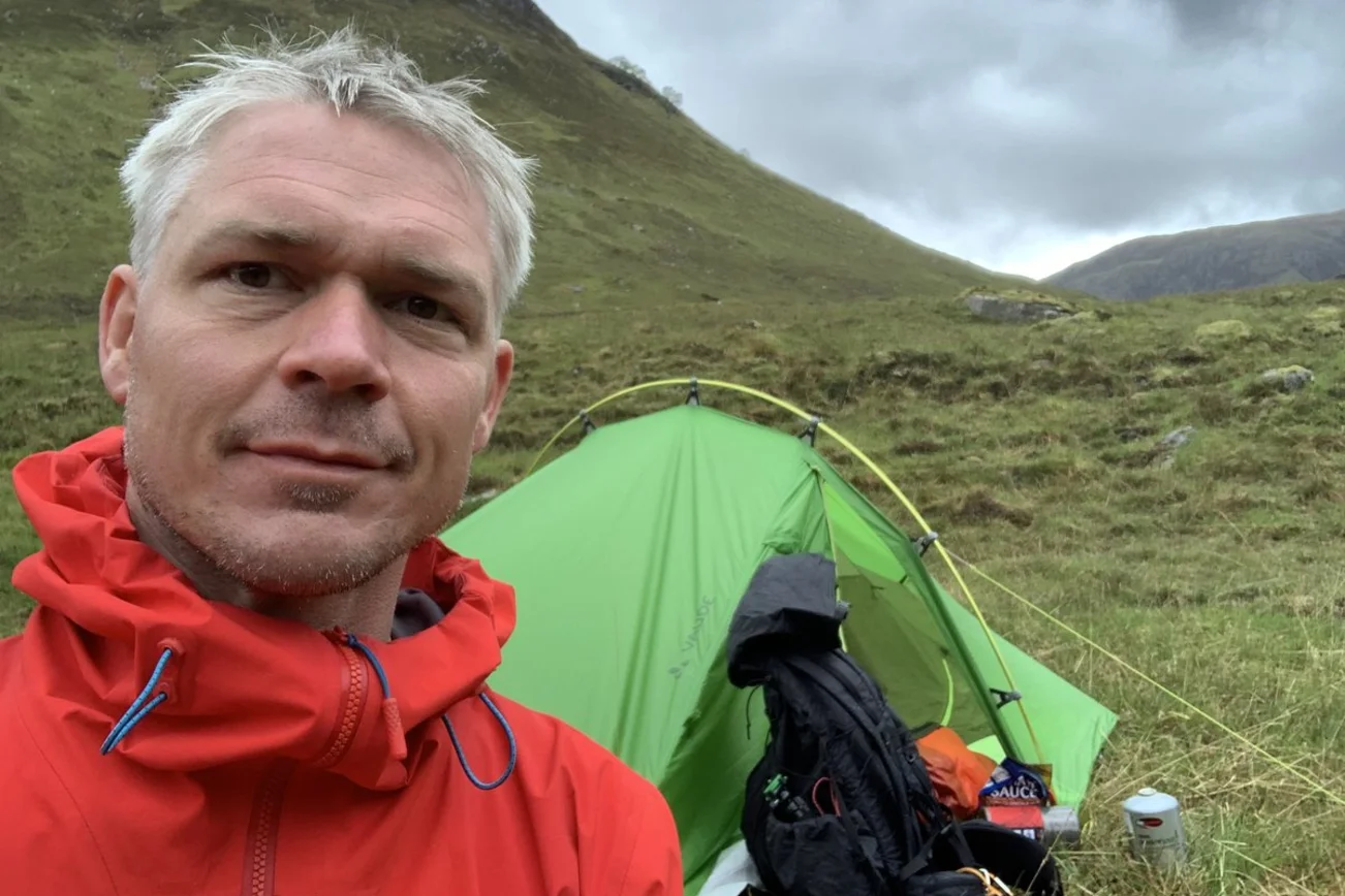 Mann mit Regenjacke vor einem Zelt in den Bergen guckt in die Kamera.