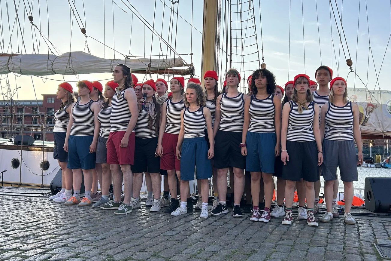 Eine Gruppe junger Menschen steht eng zusammen vor einem Segelschiff.