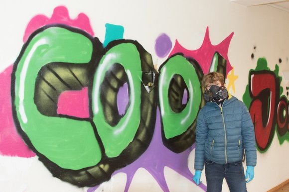 Eine junge Person mit Atemschutzmaske stehl vor einem grünen Graffiti-Schriftzug.
