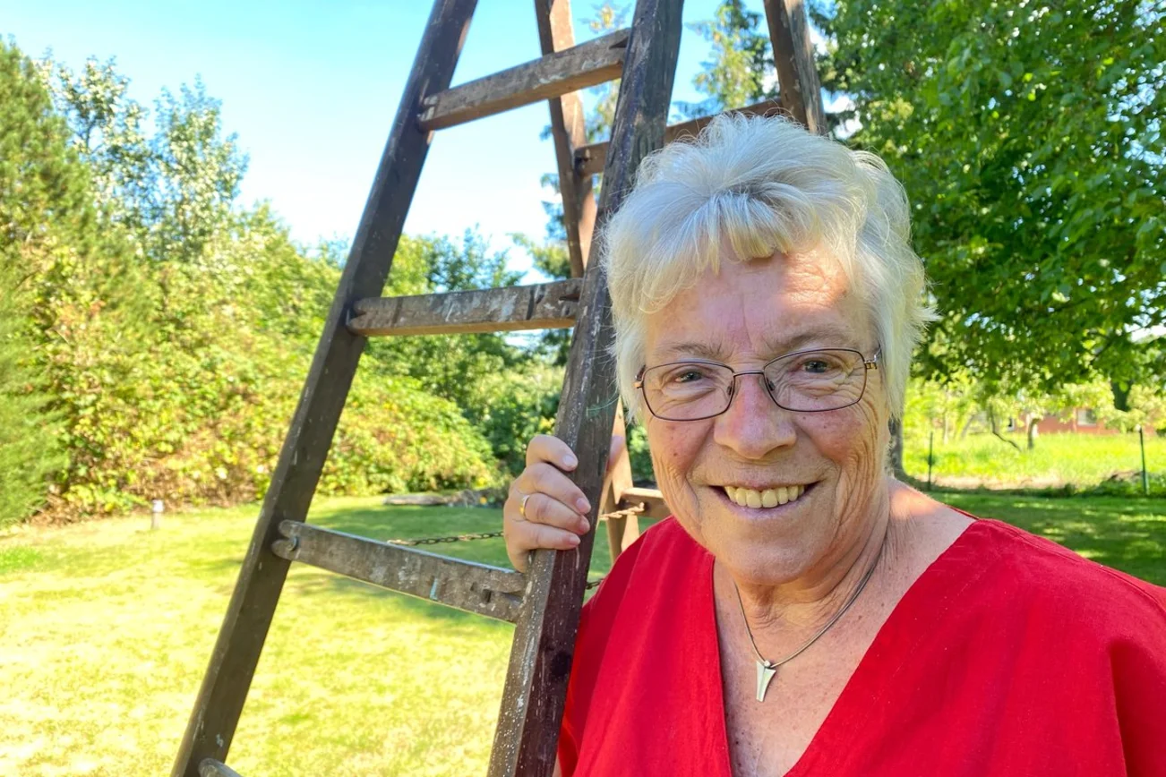 Eine Frau steht lächelnd an einer Leiter in ihrem Garten. 