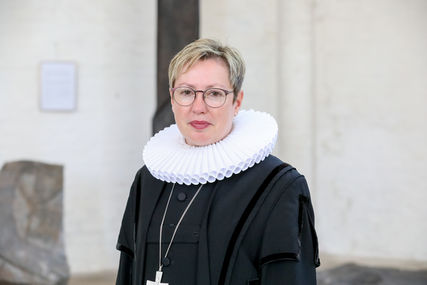 Pröpstin Petra Kallis im Talar und mit weißer Halskrause, so wie einer Kette mit goldenen Kreuz um den Hals, steht in einer Kirche und blickt in die Kamera - Copyright: Thomas Berg