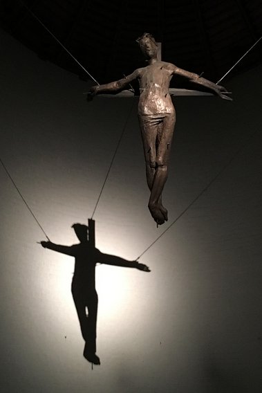 Im Altarraum hängt der gekreuzigte Christus, ein Licht erzeugt seinen Schatten auf der hinteren Wand des Altarraumessein Schatten 