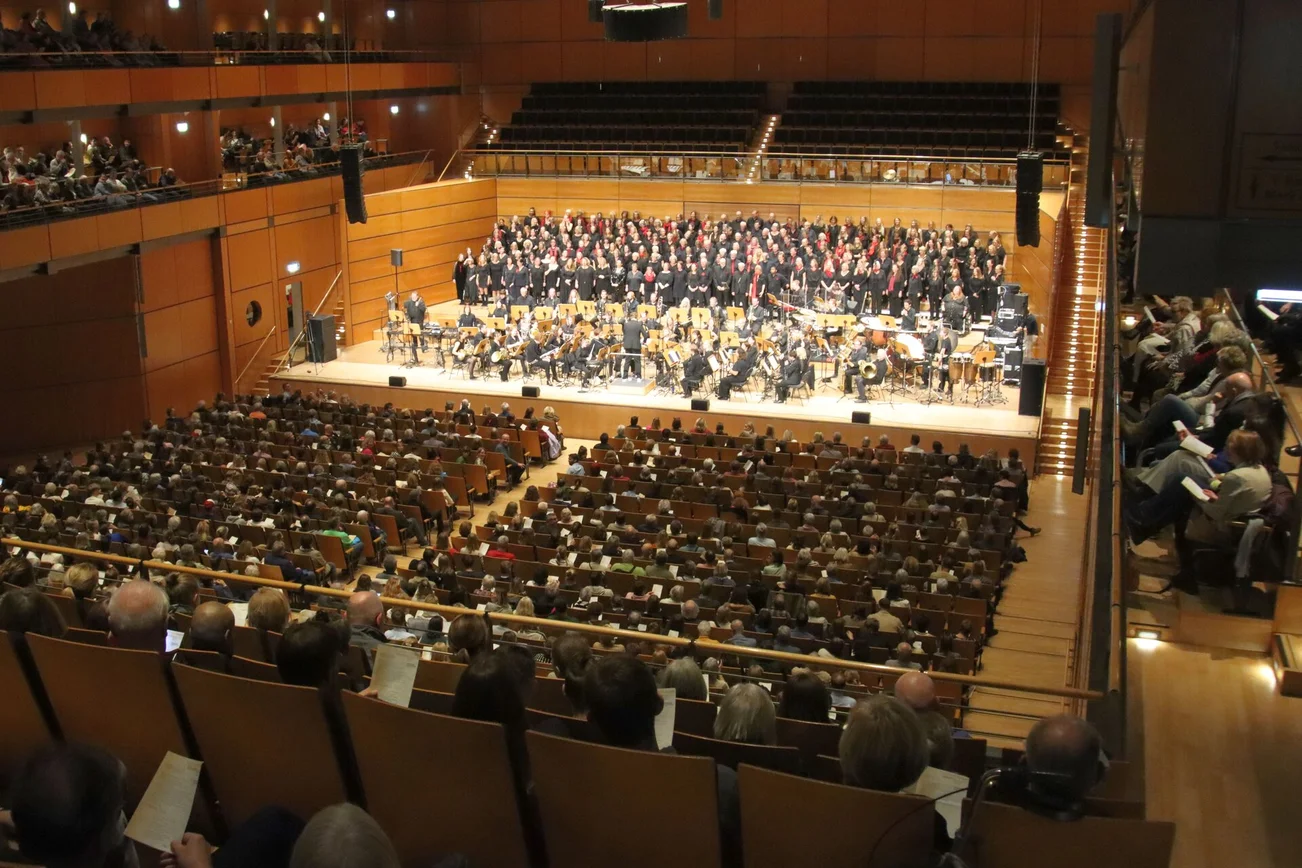 Blick in einen großen Konzertsaal auf eine voll besetzte Bühne mit Orchester und Chor.