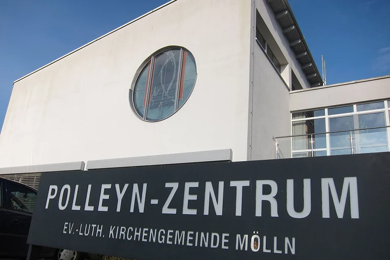 Ein großes schwarzes Schild mit weißer Schrift Polleyn-Zentrum Mölln steht vor dem Jochim-Polleyn-Haus Mölln. Es ist weiß und hat ein buntes rundes Glasfenster.
