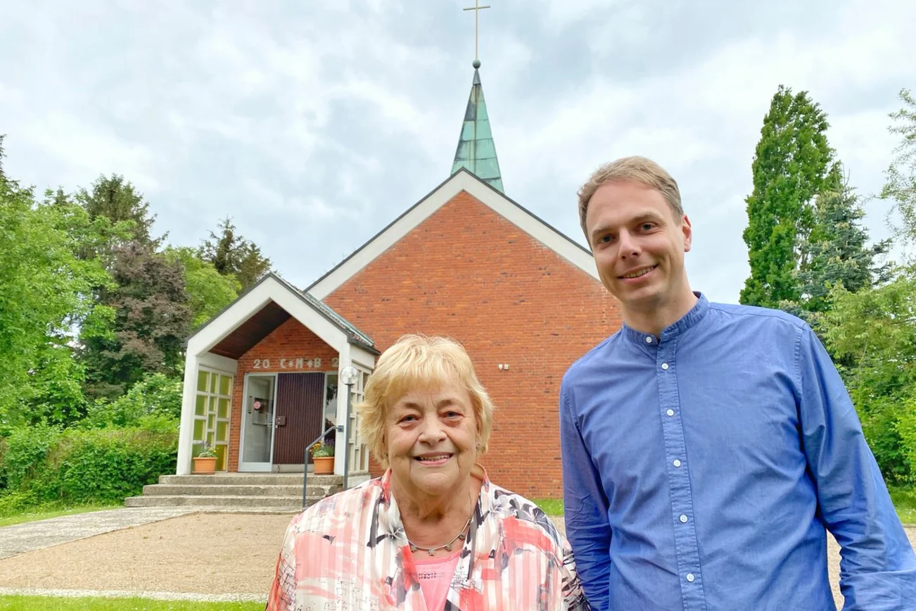 Zwei Personen stehen vor einer Kapelle und lächeln in die Kamera.
