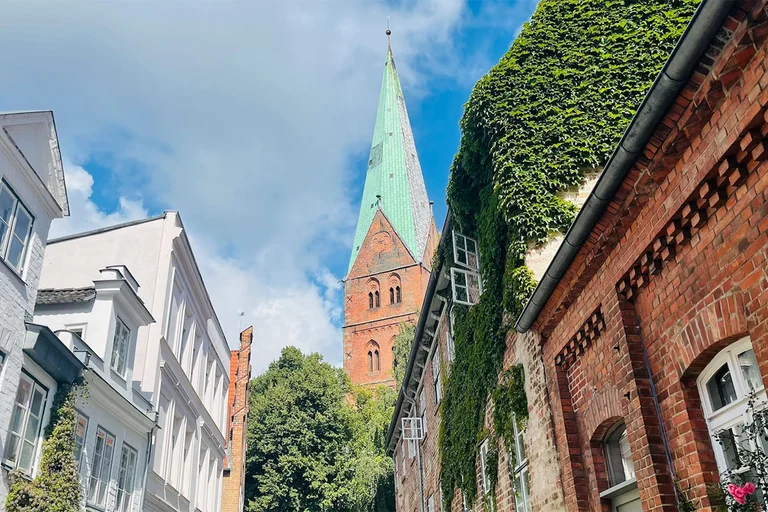 Im Hintergrund ragt der Turm von St. Aegidien in den blauen Himmel, welcher mit weißen Schleierwolken verhangen ist. Im Vordergrund die Häuserfronten der Weberstraße