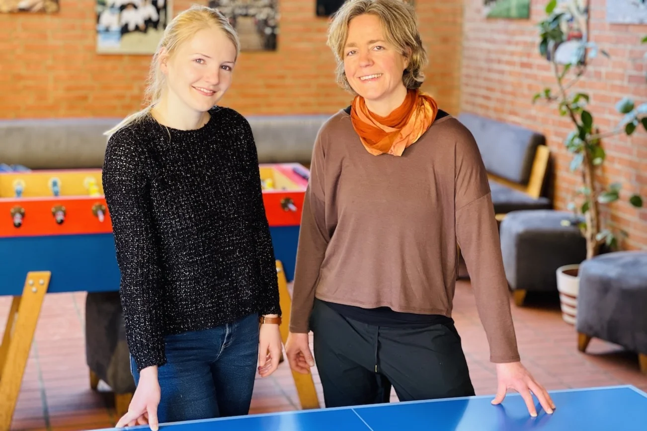 Zwei Frauen - links Merle Hillmer, Leitung der Knabenkantorei an St. Marien, und rechts die neue Chormanagerin Friederike Laukemper. Beide stehen an einer Tischtennisplatte im Probenheim und lächeln freundlich. 