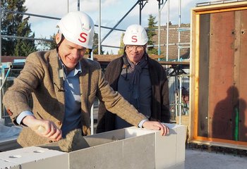 Zwei Männer mit weißen Bauhelmen stehen an deiner weißen Mauer. Der linke trägt mit einer Maurerkelle Mörtel auf. - Copyright: Annkathrin Bornholdt