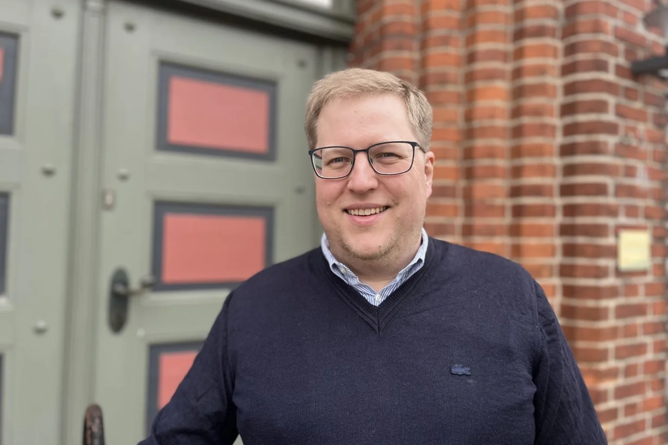 Ein Mann mit dunklem Pullover, Brille und blonden Haaren lächelt in die Kamera
