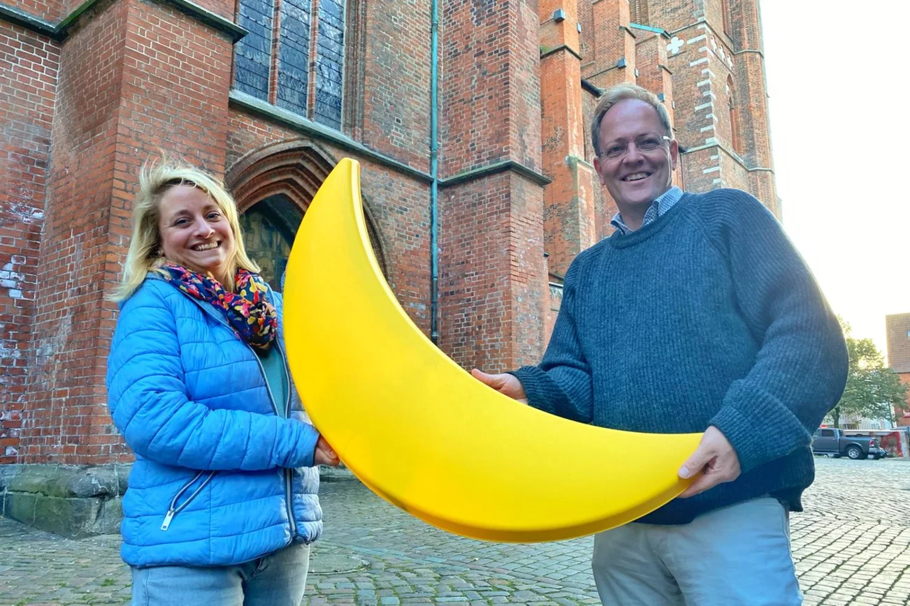 Zwei Personen stehen vor einer Kirche und halten einen Sichelnd in den Händen