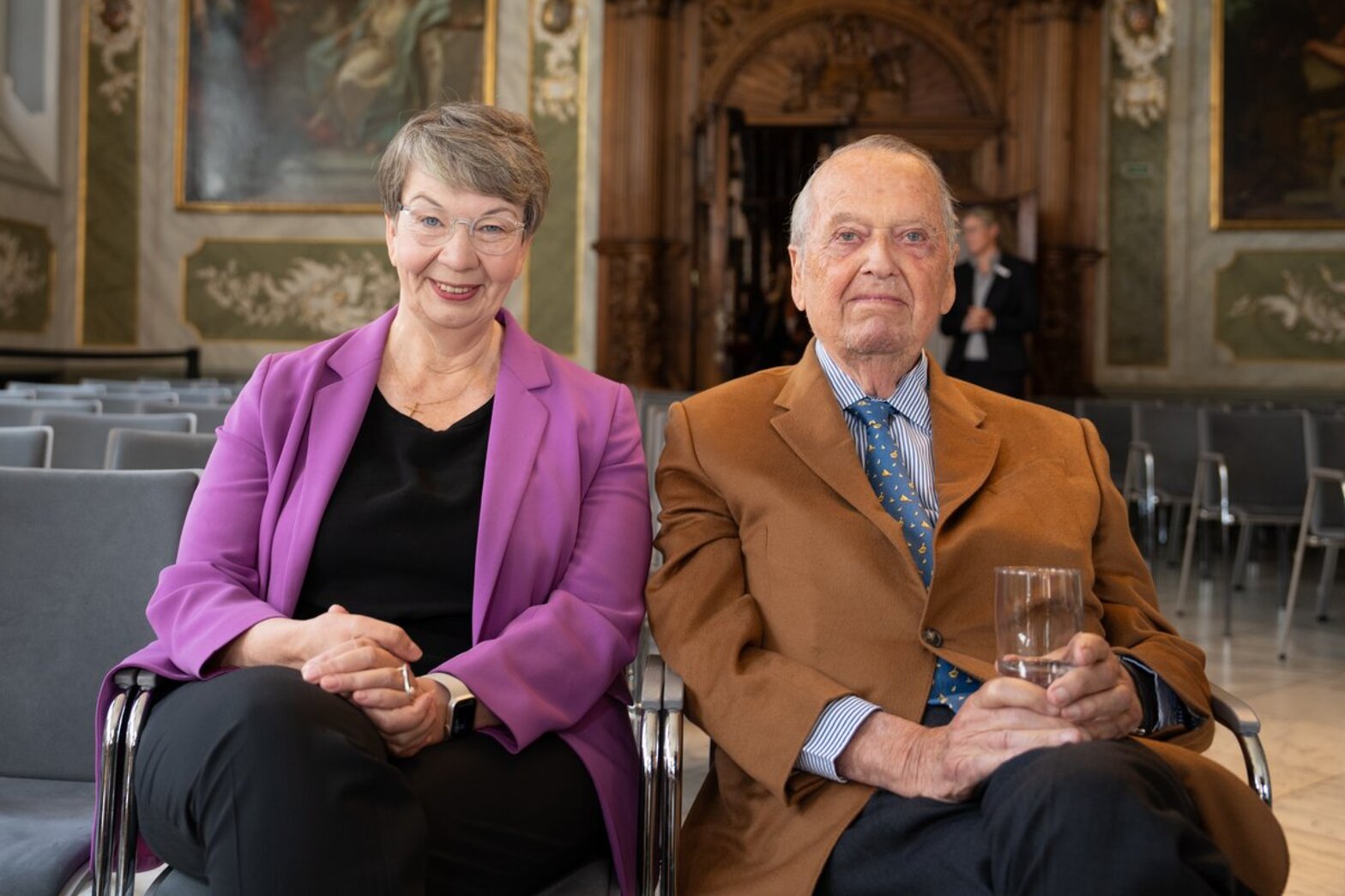 Eine Frau links und ein Mann rechts von ihr sitzen auf Stühlen, der Mann hält ein Wasserglad in den Händen.