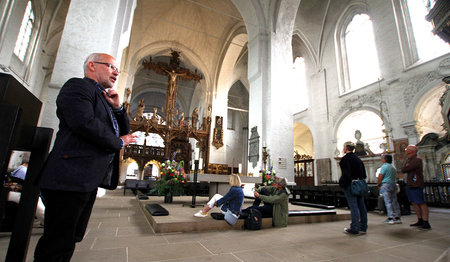 Innenraum des Dom zu Lübeck. Links im Bild steht der Pastor der Gemeinde Martin Klatt erzählt den Leuten etwas über das Triumphkreuz welches im hinteren Teil des Bildes zu sehen ist. Zwei personen sitzen am Altar auf dem Boden, drei Personen stehen rechts und schauen aufs Triumpgkreuz