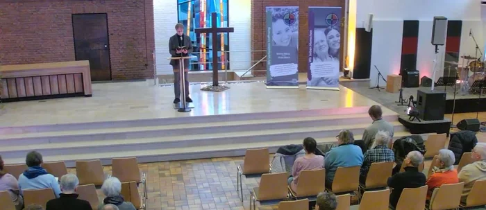 Ein Mann steht in einer Kirche vor einem Pult, vor ihm sitzen Personen auf Stühlen