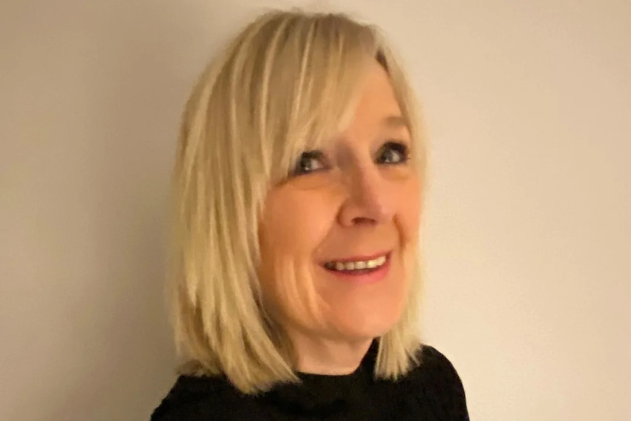 Eine blonde Frau mit einem schwarzen Oberteil lacht fröhlich in die Kamera.