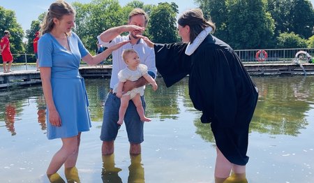 Eine Pastorin tauft ein kleines Mädchen im Krähenteich. Der Vater hält es hoch, die Mutter schaut zu freut sich. Beide Eltern halten ihre Hände als Segen über den Kopf des Kindes. 