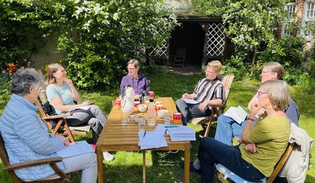 Fünf Pastorinnen und ein Vikar sitzen im sonnigen Garten um einen Tisch und bereiten das Tauffest vor.