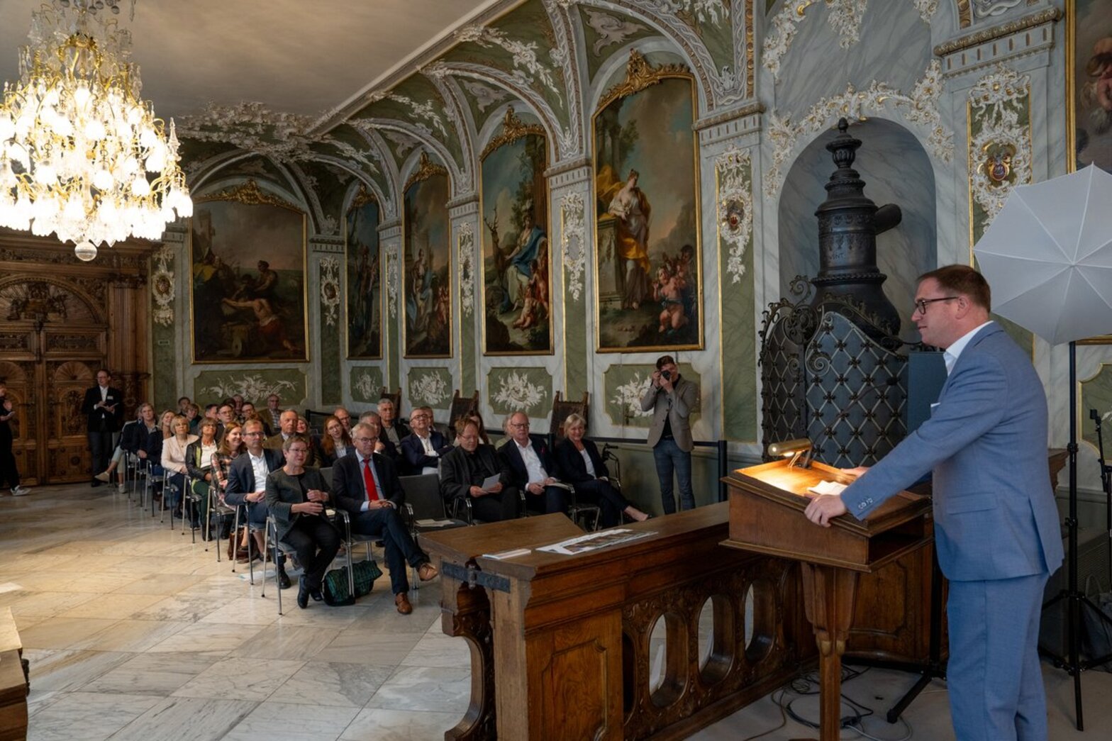 Ein Redner steht hinter einem Rednerpult, vor ihm mehrere Sitzreihen mit Menschen. Der Saal ist sehr festlich und im barocken Stil, ein Kronleuchter hängt von der Decke.