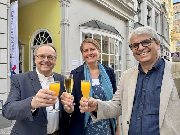 Drei Personen stoßen vor einem haus mit Getränken an. 