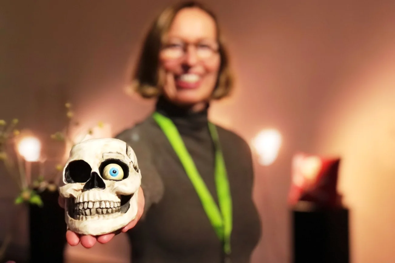 Im Vordergrund ein weißer Totenschädel. Im linken Auge liegt ein Auge aus Gips mit blauer Iris. Im Hintergrund ist unscharf die Kuratorin der Ausstellung, Anja Nitz, zu sehen.
