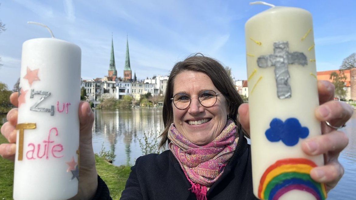 Eine Frau steht vor einem Gewässer und hält zwei Kerzen in die Kamera. 