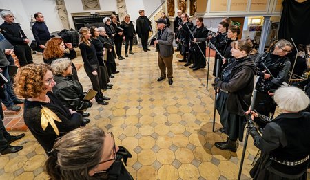 Eine Gruppe Schauspielerinnen und Schauspieler in schwarzen Uniformen steht ovalförmig in einer Kirche zusammen. 