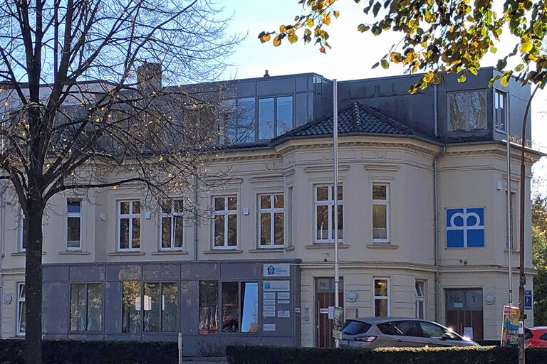 Das schöne beige Altbau-Gebäude der Gemeindediakonie. Im unteren Bereich ist eine Fensterfront zu sehen. Im Vordergrund ein Baum mit wenigen Blättern 