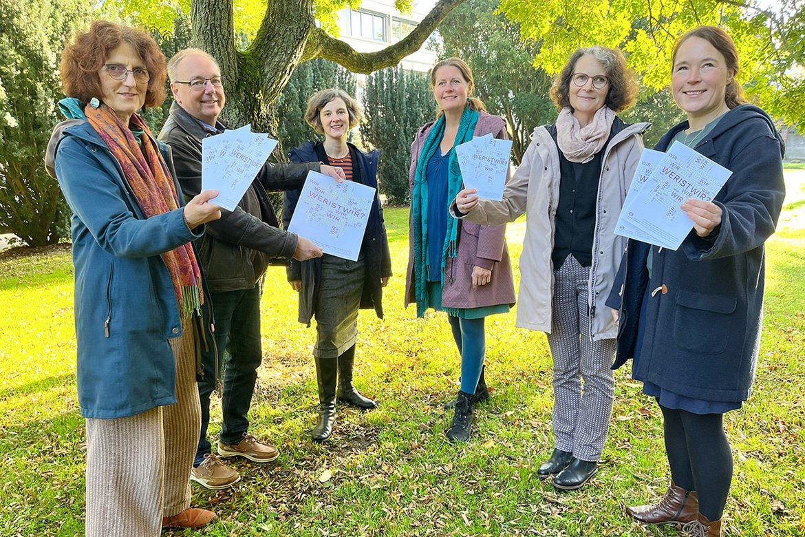 Fünf Personen stehen im Grünen im Kreis und halten die Broschüre "Wer ist wir?" in den Händen
