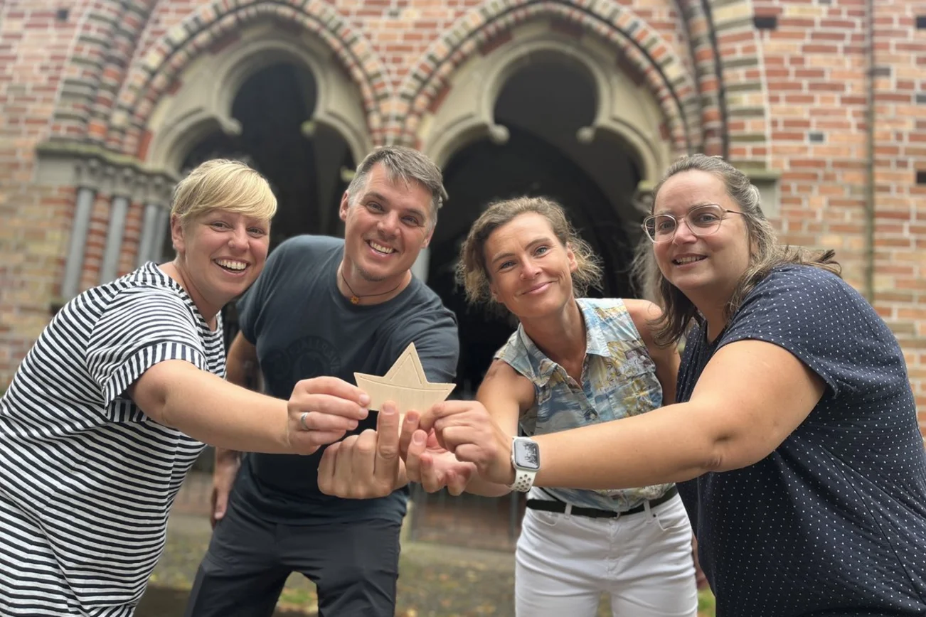 Drei Frauen und ein Mann stehen vor einer Backsteinkirche und halten ein Holzschiff hoch.
