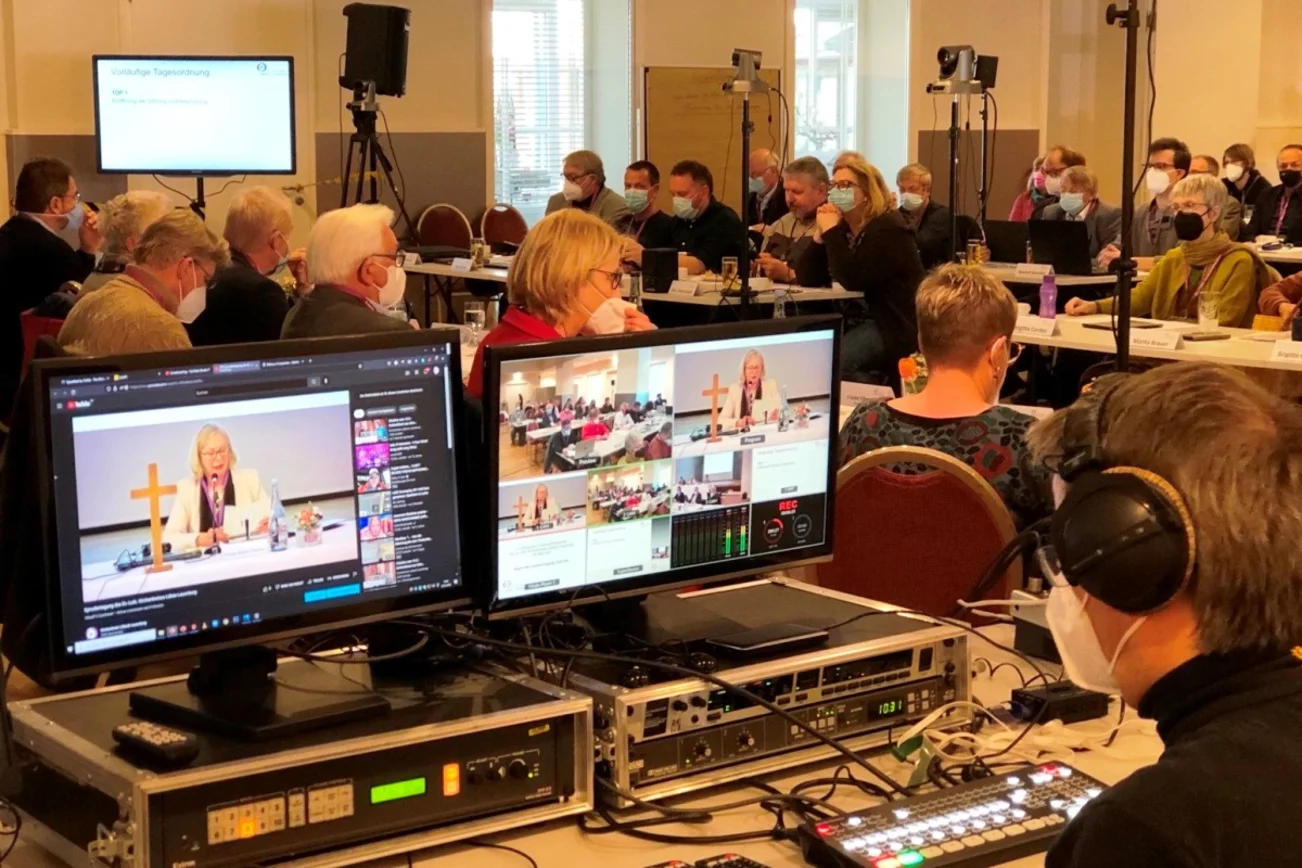 Blick in einen Saal. Menschen sitzen an Tischen. Sie tragen Masken. Im Vordergrund zwei Bildschirme mit verschiedenen Kameraeinstelllungen. 