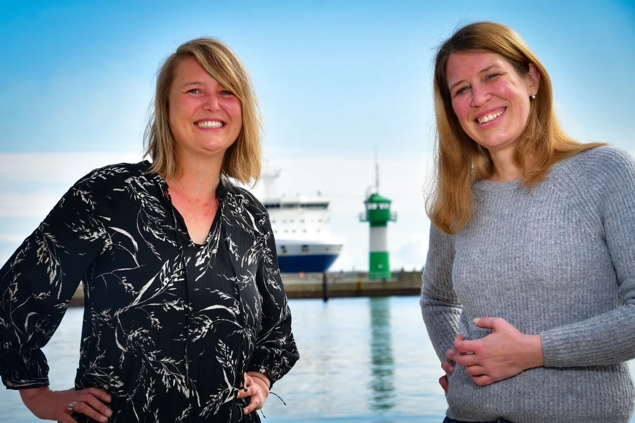 Zwei Frauen blicken freundlich in die Kamera, im Hintergrund die die Nordermole in Travemünde: Grün-weißer Leuchtturm, der Bug eines Containerschiffs