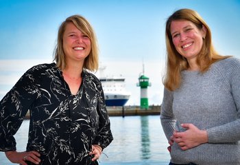 Zwei Frauen blicken freundlich in die Kamera, im Hintergrund die die Nordermole in Travemünde: Grün-weißer Leuchtturm, der Bug eines Containerschiffs - Copyright: Bastian Modrow