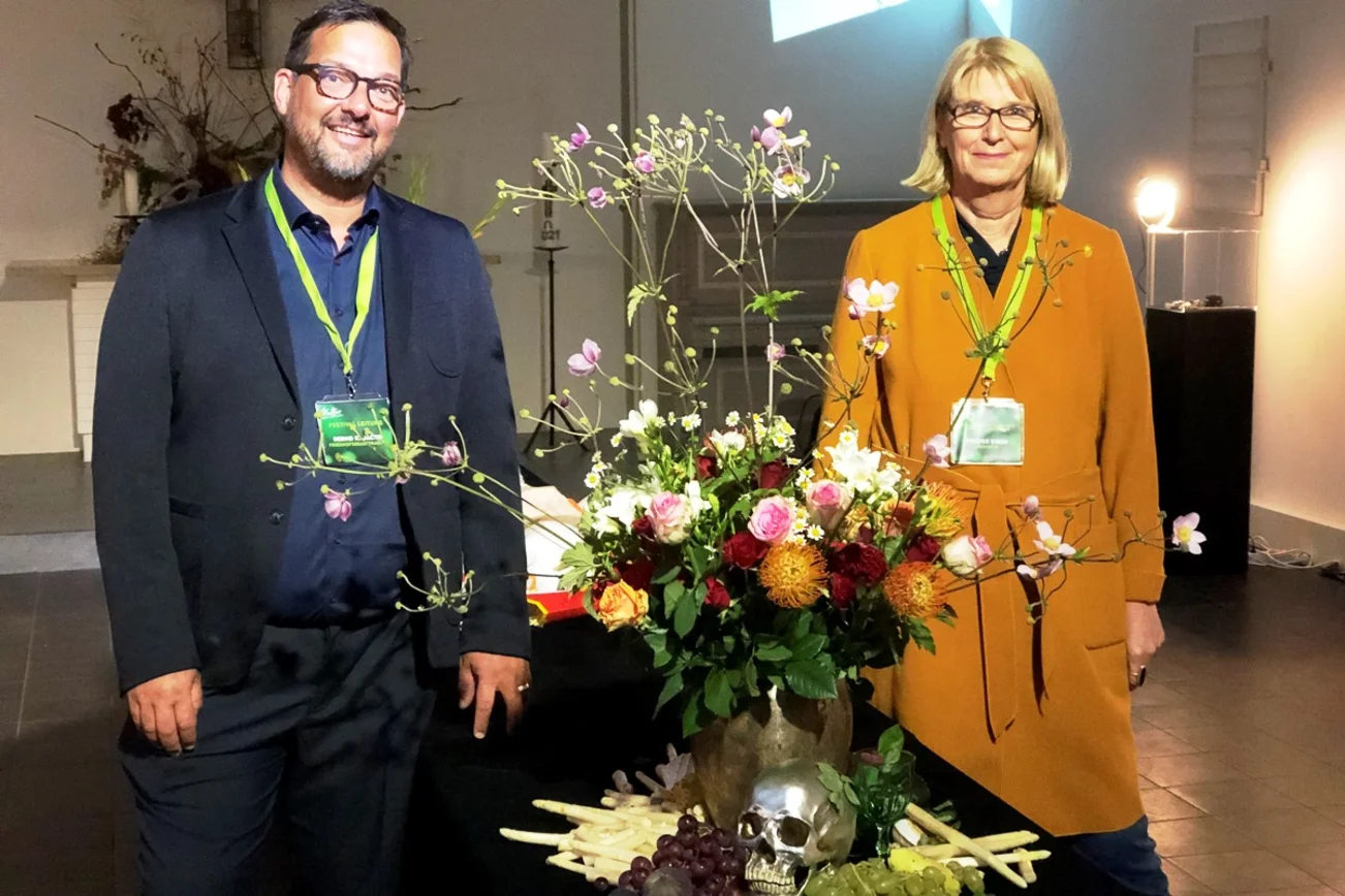 Ein Mann und eine Frau stehen rechts und links eines Blumengestecks. Am Fuß der Vase liegen Trauben, ein silberner Totenkopf. 