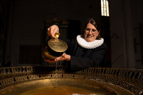 Eine Frau im schwarzen Talar und weißer Halskrause steht hinter einem goldenen Taufbeckenrand und hält ein goldenes Gefäß über die Taufschale. Die Frau trägt eine Brille und das mittellange braune Haar zurückgebunden.