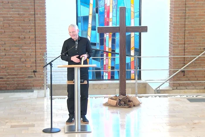 Ein Mann mit Brille und grauen Haaren steht hinter einem Stehpult und hält ein Buch. Im Hintergrund steht ein Holzkreuz und ein buntes Kirchenfenster ist zu sehen