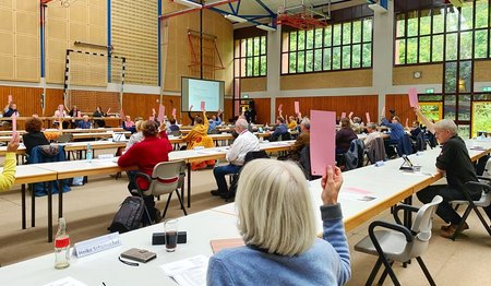 Der Blick in eine Sporthalle, die mit Tischen und Stühlen ausgestattet ist. Im Vordergrund die Rückansicht einer Frau. Sie sitzt an einem Tisch, trägt eine hellblaue Jacke und hebt eine rote Abstimmungskarte. 